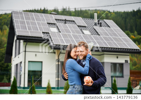 Happy couple hugging and holding piggy bank with cash near house with solar panels. Financial savings from renewable energy and eco home lifestyle concept 143779136