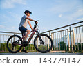 Man riding electric mountain bike on city promenade, side view cyclist against urban skyline and blue sky, active lifestyle and eco transport concept 143779147