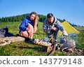 Man and woman by campfire in mountain meadow. Female tourist holding cup while husband tending to kettle on fire. Yellow tent and lush green hills in background, all under clear blue sky. 143779148