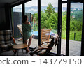 Woman enjoying quiet morning by panoramic window in a modern mountain cabin, admiring green hillside scenery, terrace lounge area and outdoor hot tub 143779150