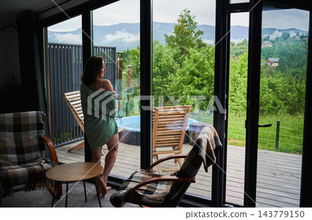 Woman enjoying quiet morning by panoramic window in a modern mountain cabin, admiring green hillside scenery, terrace lounge area and outdoor hot tub Woman enjoying quiet morning by panoramic window in a modern mountain cabin, admiring green hillside scenery, terrace lounge area and outdoor hot tub 143779150