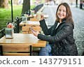 Portrait of young woman smiling at camera while holding latte and smartphone at outdoor cafe, sitting at wooden table, wearing casual jacket, enjoying leisure time near green park 143779151