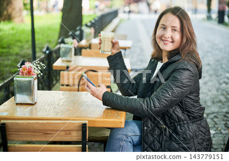 Portrait of young woman smiling at camera while holding latte and smartphone at outdoor cafe, sitting at wooden table, wearing casual jacket, enjoying leisure time near green park 143779151