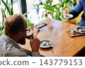 Man drinking cappuccino at cafe table during conversation, relaxed break with colleague, cozy atmosphere with smartphone, dessert plate and indoor plants in soft daylight 143779153