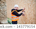 Construction worker in safety vest and helmet fixing OSB panels to exterior wall with cordless drill, illustrating facade installation, site safety and modern house building process 143779154