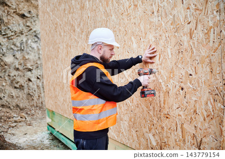 Construction worker in safety vest and helmet fixing OSB panels to exterior wall with cordless drill, illustrating facade installation, site safety and modern house building process Construction worker in safety vest and helmet fixing OSB panels to exterior wall with cordless drill, illustrating facade installation, site safety and modern house building process 143779154