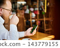 Man drinking cappuccino and checking smartphone with green screen in cozy cafe, side view through glass, warm sunlight, relaxed lifestyle, mobile browsing and digital communication concept 143779155