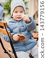Toddler boy eating cookie while sitting on chair outdoors. Wearing warm jacket and hat on terrace in autumn 143779156