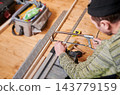 Man cutting metal profile with hacksaw, close up of hands during construction work. Indoor workshop with tools, measuring tape and materials, DIY renovation, craftsmanship concept 143779159