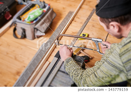Man cutting metal profile with hacksaw, close up of hands during construction work. Indoor workshop with tools, measuring tape and materials, DIY renovation, craftsmanship concept Man cutting metal profile with hacksaw, close up of hands during construction work. Indoor workshop with tools, measuring tape and materials, DIY renovation, craftsmanship concept 143779159