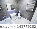 Modern bathroom interior with sink cabinet, toilet and heated towel rail. Stylish washroom with dark textured walls, marble floor tiles, backlit mirror and bathtub edge 143779162