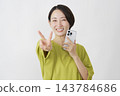 一位中年婦女在白色背景前舉起智慧型手機，比出和平手勢。 143784686
