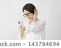 一位中年婦女雙手抱頭，在白色背景下看著她的智慧型手機。 143784694