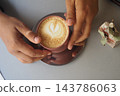 Hands holding a cup of coffee with latte art at a table 143786063