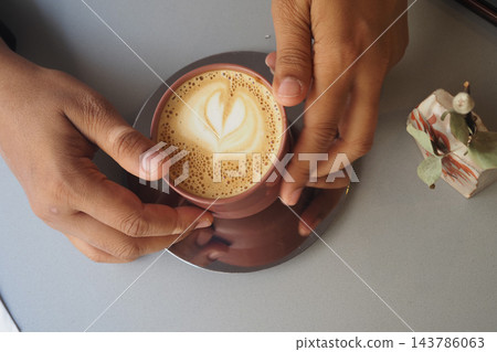 Hands holding a cup of coffee with latte art at a table Hands holding a cup of coffee with latte art at a table 143786063