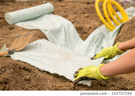 Hands are carefully applying protective fabric to well prepared soil in a landscaping trench, ensuring effective erosion prevention and enhancing the durability of the installation. Hands are carefully applying protective fabric to well prepared soil in a landscaping trench, ensuring effective erosion prevention and enhancing the durability of the installation. 143787589