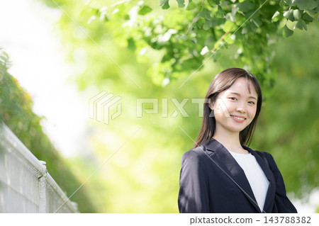 一位年輕的日本女員工，剛畢業不久，在美麗的春日綠蔭下方帶著微笑，直視鏡頭，展現了大學生求職或新進員工的形象。 143788382