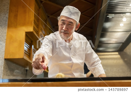一位經驗豐富的壽司師傅在吧台座位上現場製作壽司。 一位經驗豐富的壽司師傅在吧台座位上現場製作壽司。 143789897