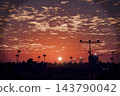 Airport runway approach lights at sunset, sun on horizon, palm trees and city silhouette under orange sky with clouds, 3D illustration of an airfield in warm evening light 143790042