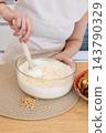 Kneading dough. Culinary work. Female baker hand mixing wet flour ingredient in glass bowl preparing healthy cookies with pine nuts. 143790329