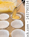 Preparing muffin. Treat recipe. Spoon pouring sweet yellow batter in paper liners baking tin on home kitchen counter with dough bowl. 143790334