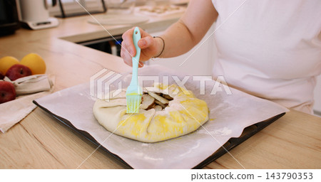 Sweet tart. Glazing pie crust. Woman baker hand brushing round galette with liquid yellow egg wash on baking sheet at home kitchen. Sweet tart. Glazing pie crust. Woman baker hand brushing round galette with liquid yellow egg wash on baking sheet at home kitchen. 143790353