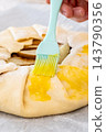 Sweet tart. Glazing pie crust. Woman baker hand brushing round galette with liquid yellow egg wash on baking sheet at home kitchen. 143790356