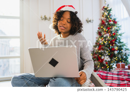 Merry Christmas. African woman in Santa hat having video chat in laptop. Blogger in festive mood recording video in social network. Girl having video call with friends family celebrating Christmas Merry Christmas. African woman in Santa hat having video chat in laptop. Blogger in festive mood recording video in social network. Girl having video call with friends family celebrating Christmas 143790425
