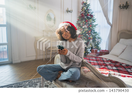 African American woman holding smartphone scrolling typing shopping online buying Christmas gifts. Girl buying on Internet chatting in social network enjoying Christmas winter holidays at home 143790430