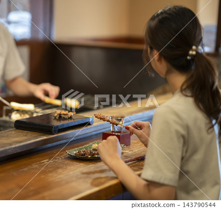 一位年輕女子獨自一人在烤雞串店的櫃檯前吃喝,背影清晰可見。 一位年輕女子獨自一人在烤雞串店的櫃檯前吃喝,背影清晰可見。 143790544