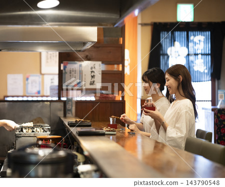 兩位年輕女子在吧台座位上享用烤雞串和清酒。 兩位年輕女子在吧台座位上享用烤雞串和清酒。 143790548