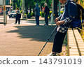 A senior woman sits on a block of nearby plants while waiting for the traffic light to go off at a crosswalk. 143791019