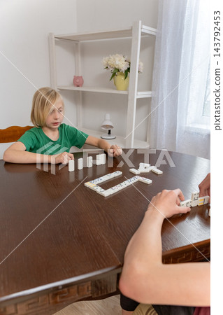 Young boy intently playing dominoes with another person at a wooden table indoors, focused on placing tiles in a quiet family moment of learning, strategy and bonding 143792453