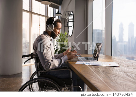 Businessman in wheelchair in headphones, engaged in telemeeting using laptop Businessman in wheelchair in headphones, engaged in telemeeting using laptop 143793424