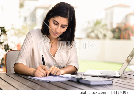 Woman taking notes in notebook sitting at table in courtyard Woman taking notes in notebook sitting at table in courtyard 143793953