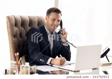 Smiling businessman in suit talking on landline phone at workplace 143793972
