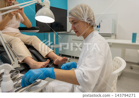 Female podiatrist examines patients feet at clinic, holding small instrument beside gloved hands. Focused foot care professional works under bright lamp in clean treatment room. 143794271