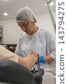 Focused nail technician cleans client toenails during beauty salon pedicure appointment. Aesthetician dressed in white uniform applies skincare cosmetics, showing serious professional concentration. 143794275