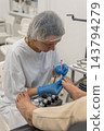 Skilled esthetician performs detailed pedicure procedure on customer feet using specialized precision tool. Dedicated worker carefully tends to toenail hygiene inside bright clinical clinic. 143794279