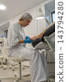 Foot care specialist examines patients foot under bright clinic lamp, holding toes during nail treatment. Attentive podiatrist wears blue gloves and protective cap beside treatment chair. 143794280