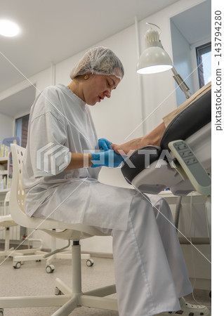 Foot care specialist examines patients foot under bright clinic lamp, holding toes during nail treatment. Attentive podiatrist wears blue gloves and protective cap beside treatment chair. 143794280