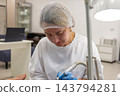 Female foot care specialist files patients toenail with electric podiatry tool. Focused practitioner wears gloves and hair cover while providing treatment in clean clinic. 143794281