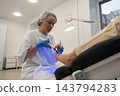 Podiatrist applies blue light to patients toenail while guiding small instrument. Attentive clinician works in clean treatment room with gloves and hair cover, positioning instrument at nail. 143794283