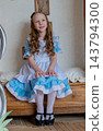 Smiling Girl Seated On Bench Wearing Blue Ruffled Dress, Hands Folded On Lap, Polished Mary Jane Shoes, Soft Curls, Warm Natural Light, Neutral Wallpaper Backdrop, Gentle Confident Expression. 143794300