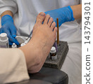 Pedicurist wearing protective gloves treats human foot during clinical pedicure session. Carefully arranged bottles and treatment brushes support thorough toenail health procedure. 143794301