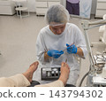 Careful specialist delivers pedicure treatment to person resting feet in chair, inspecting toenail condition while working with fine medical tools during clinical hygiene appointment. 143794302