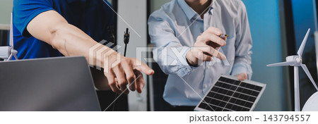 Two Asian engineers wearing hardhats and safety vests discussing solar panel installation and renewable energy project in the office. 143794557