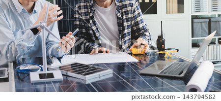 Two Asian engineers wearing hardhats and safety vests discussing solar panel installation and renewable energy project in the office. Two Asian engineers wearing hardhats and safety vests discussing solar panel installation and renewable energy project in the office. 143794582