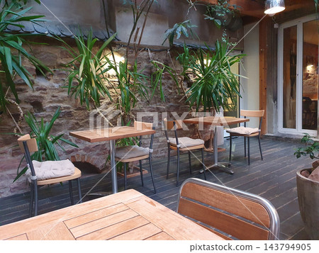 A cozy indoor cafe patio featuring wooden tables, cushioned chairs, and lush green plants against a rustic stone wall. Soft lighting and a warm, relaxed atmosphere perfect for conversation or quiet 143794905