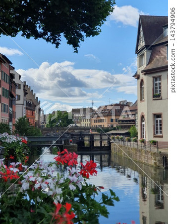 Picturesque Petite France canal scene in Strasbourg with colorful flower boxes, historic timbered houses, pedestrian bridge and calm water reflections under a bright blue summer sky. Charming Picturesque Petite France canal scene in Strasbourg with colorful flower boxes, historic timbered houses, pedestrian bridge and calm water reflections under a bright blue summer sky. Charming 143794906